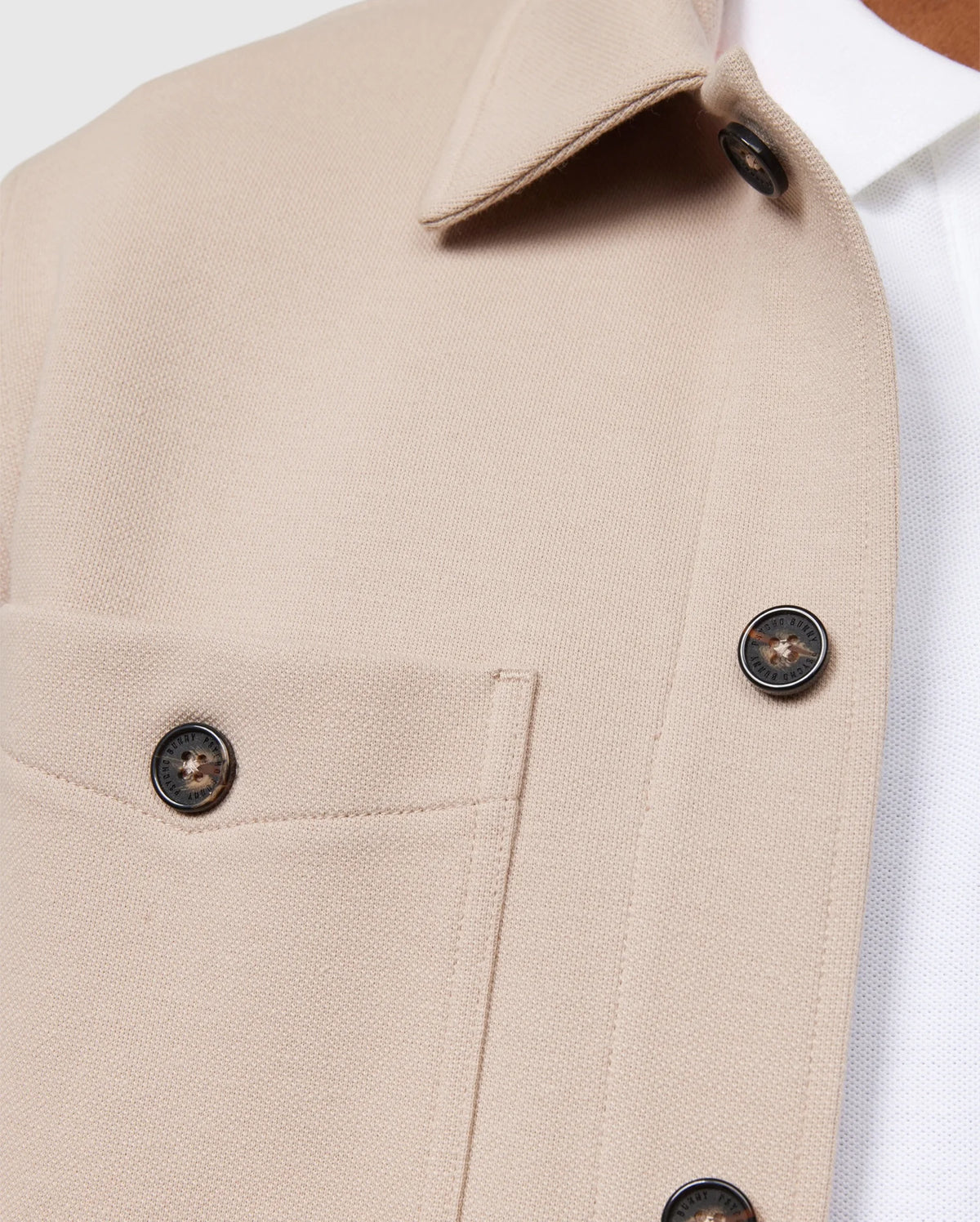 Close-up of the Psycho Bunny MENS BEDFORD SHIRT JACKET - B6J290B200 in beige, featuring a chest pocket and dark buttons. The jacket, worn over a white collared shirt, showcases the fabric texture and button details. This shot highlights the elevated style and exceptional on-body feel of the upper section design and stitching.