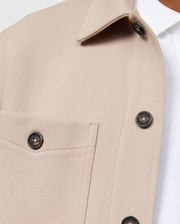 Close-up of the Psycho Bunny MENS BEDFORD SHIRT JACKET - B6J290B200 in beige, featuring a chest pocket and dark buttons. The jacket, worn over a white collared shirt, showcases the fabric texture and button details. This shot highlights the elevated style and exceptional on-body feel of the upper section design and stitching.
