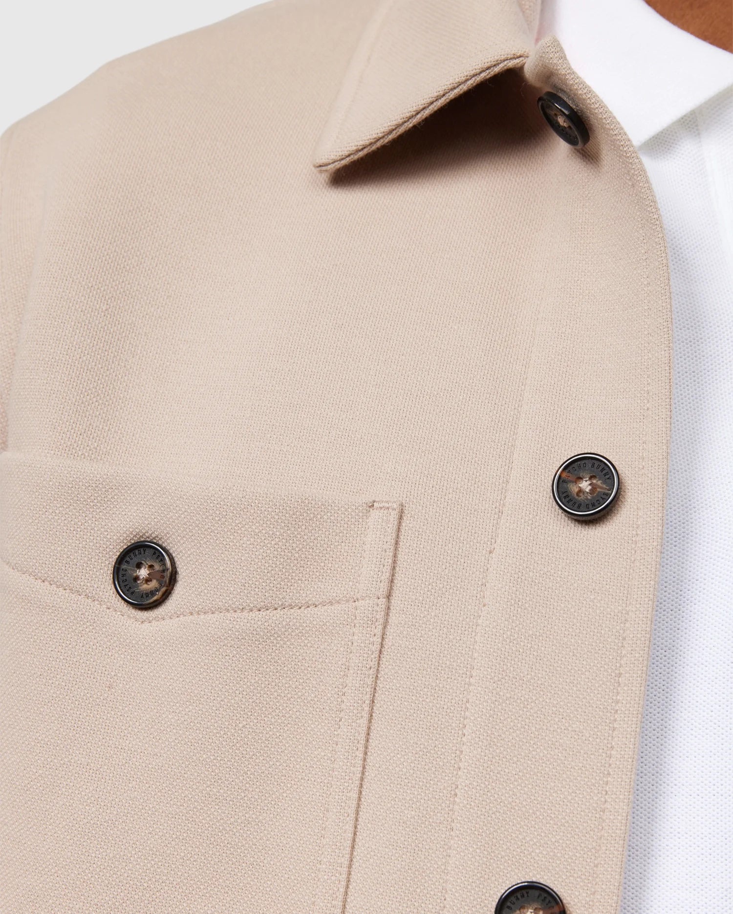 Close-up of the Psycho Bunny MENS BEDFORD SHIRT JACKET - B6J290B200 in beige, featuring a chest pocket and dark buttons. The jacket, worn over a white collared shirt, showcases the fabric texture and button details. This shot highlights the elevated style and exceptional on-body feel of the upper section design and stitching.
