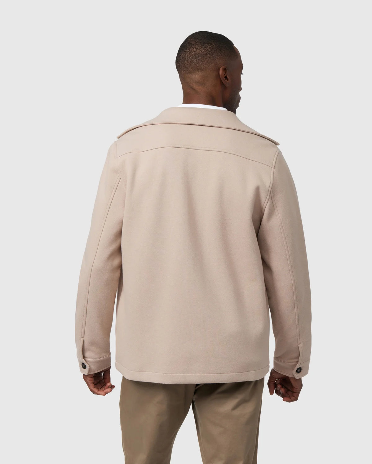 A man is standing with his back to the camera, wearing a stylish beige MENS BEDFORD SHIRT JACKET - B6J290B200 by Psycho Bunny, featuring a collar, button cuffs, and shoulder straps. He has paired it with khaki pants against a plain, light gray background.
