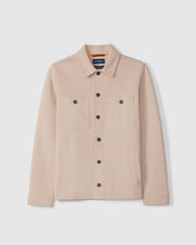 The Psycho Bunny Men's Bedford Shirt Jacket (B6J290B200) is a beige button-up jacket featuring long sleeves, two large chest pockets with buttoned flaps, and two side pockets. It has a collar and black buttons down the front. The interior boasts an orange lining near the collar with a visible brand label, ensuring an exceptional on-body feel.