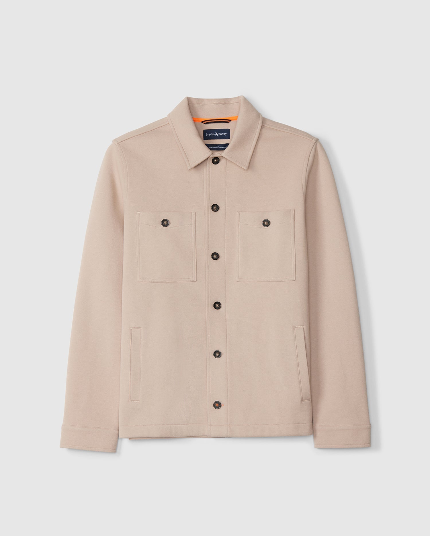 The Psycho Bunny Men's Bedford Shirt Jacket (B6J290B200) is a beige button-up jacket featuring long sleeves, two large chest pockets with buttoned flaps, and two side pockets. It has a collar and black buttons down the front. The interior boasts an orange lining near the collar with a visible brand label, ensuring an exceptional on-body feel.