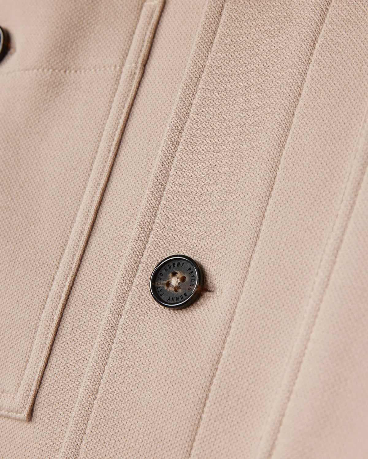 Close-up of the beige Psycho Bunny men's Bedford shirt jacket (B6J290B200) featuring two vertical seams and a round, black button inscribed "ORIGINAL STYLE." The texture of the fabric is distinct, showcasing a subtle weave pattern that enhances its elevated style and offers an exceptional on-body feel.