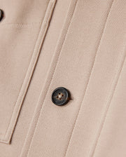 Close-up of the beige Psycho Bunny men's Bedford shirt jacket (B6J290B200) featuring two vertical seams and a round, black button inscribed "ORIGINAL STYLE." The texture of the fabric is distinct, showcasing a subtle weave pattern that enhances its elevated style and offers an exceptional on-body feel.