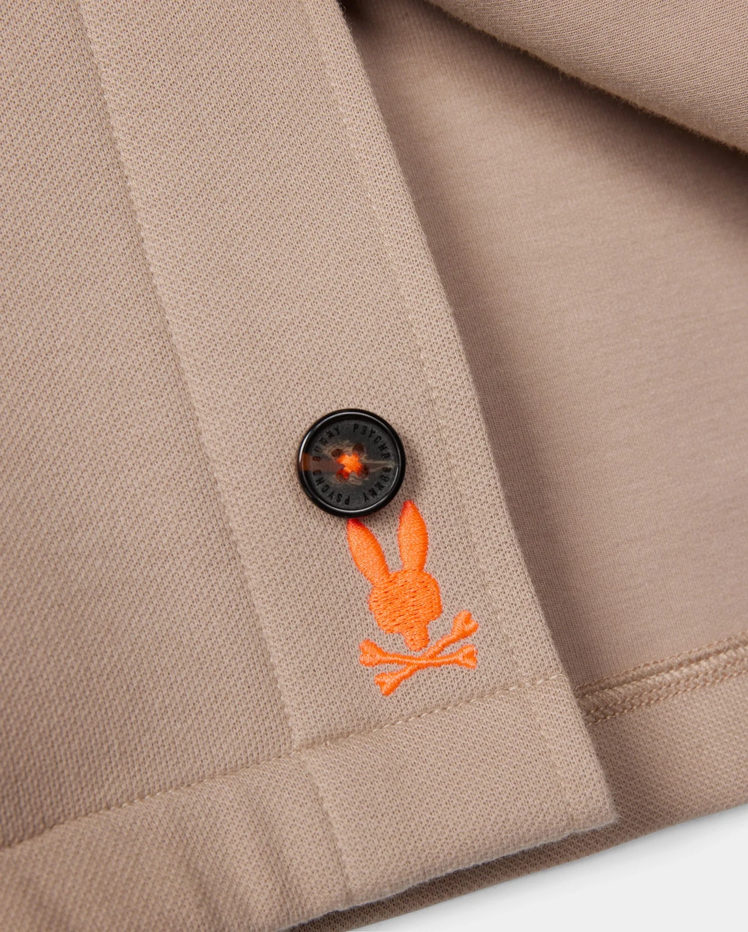 Close-up of a beige fabric with a black button stitched onto it. Below the button, there is an orange embroidered logo featuring Psycho Bunny's iconic bunny head with crossed bones underneath. The edge of the fabric is neatly hemmed, showcasing the exceptional on-body feel and elevated style of the MENS BEDFORD SHIRT JACKET - B6J290B200 by Psycho Bunny.