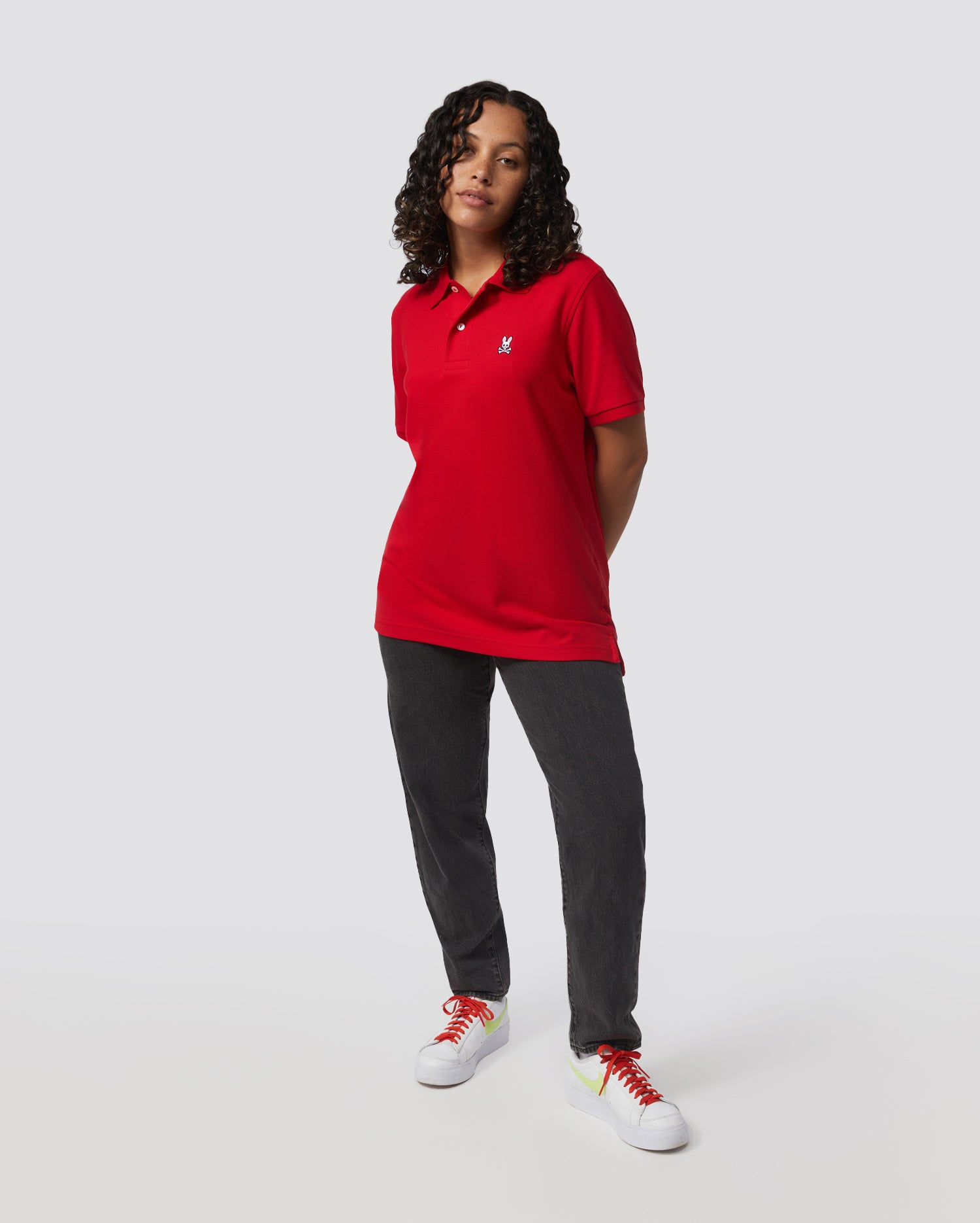 Wearing a red Psycho Bunny Men's Classic Pique Polo Shirt (B6K001CRPC) made of 100% Pima cotton with mother-of-pearl buttons, black pants, and white sneakers with colorful laces, a person stands against a gray backdrop, hands behind their back and gazing forward.