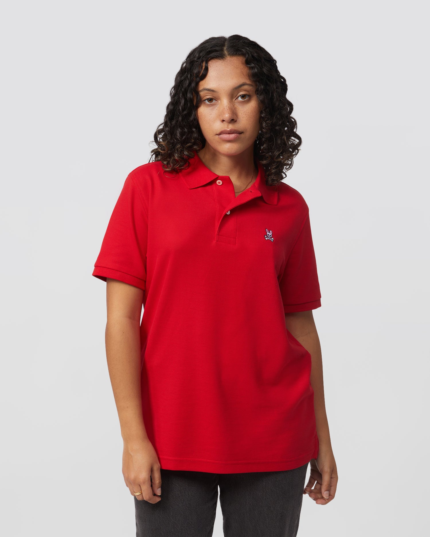 A person with curly hair is wearing a Psycho Bunny Men's Classic Pique Polo Shirt in red, made of 100% Pima cotton, paired with dark pants against a light gray background. The polo features a small embroidered logo on the left chest and mother-of-pearl buttons.