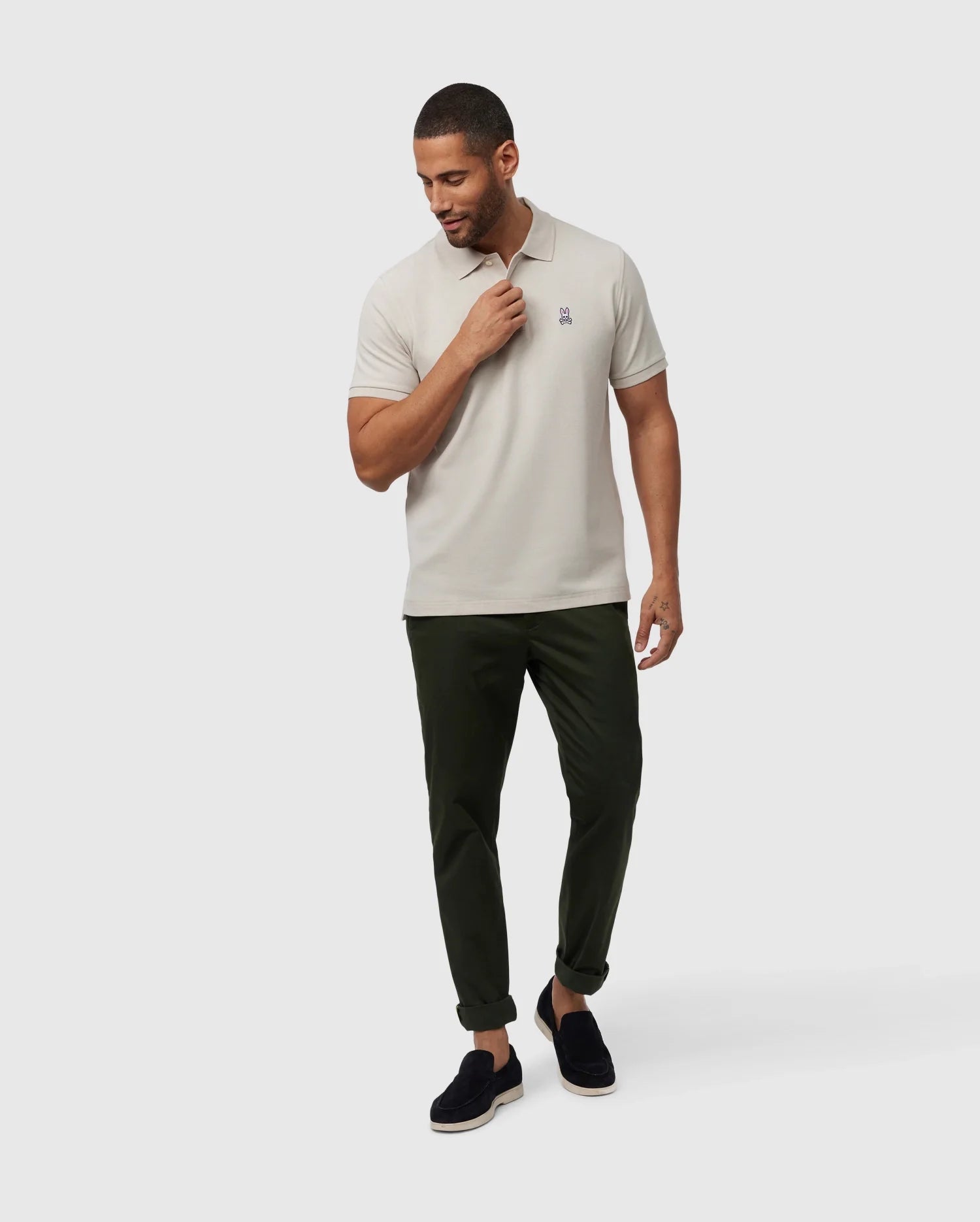 A man in a Psycho Bunny MENS CLASSIC PIQUE POLO SHIRT - B6K001B200, light beige, paired with dark green pants stands against a white background. He wears black slip-on shoes and has neat short hair. His left hand touches his neck while he gazes downwards, showcasing men's fashion.