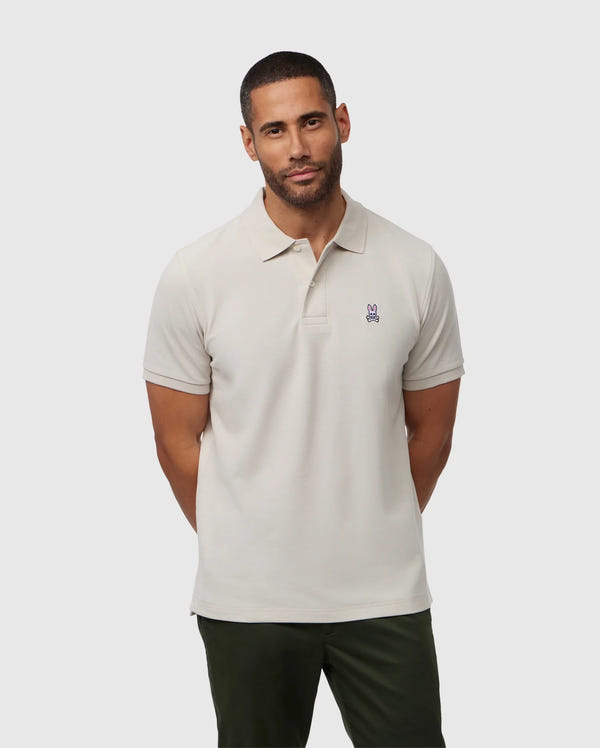 A man with short hair and a beard is wearing the Psycho Bunny Men's Classic Pique Polo Shirt (B6K001B200) made from soft Pima cotton, featuring a small bunny logo. He stands against a plain background with his hands behind his back, showcasing this staple of men's fashion.