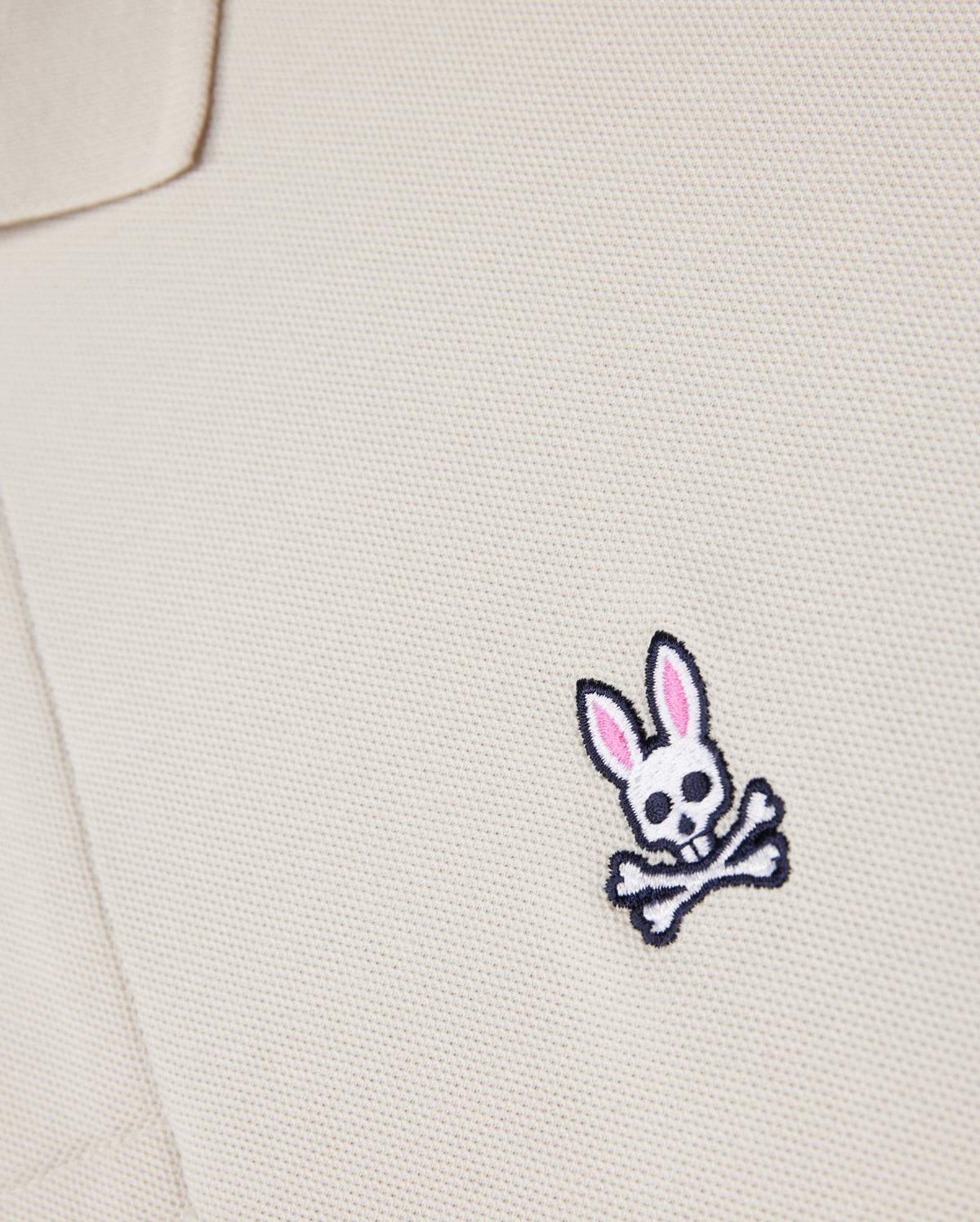 A close-up of the Psycho Bunny beige MENS CLASSIC PIQUE POLO SHIRT - B6K001B200, made from premium Pima cotton, showcases a small bunny skull emblem with pink ears and crossbones. It adds an edgy twist to men's fashion.