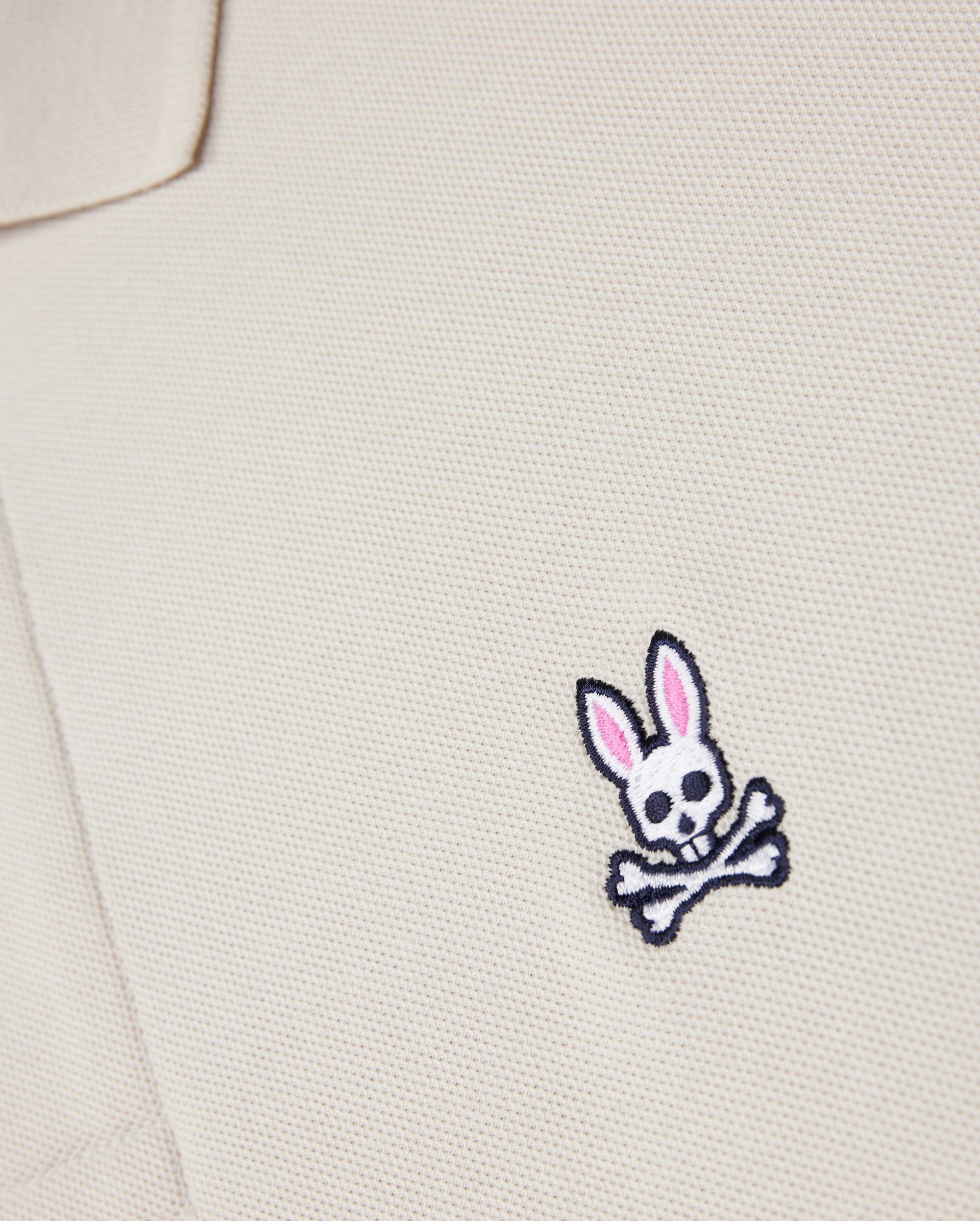 A close-up of the Psycho Bunny beige MENS CLASSIC PIQUE POLO SHIRT - B6K001B200, made from premium Pima cotton, showcases a small bunny skull emblem with pink ears and crossbones. It adds an edgy twist to men's fashion.