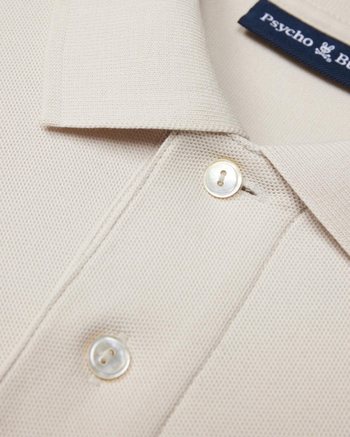 The Psycho Bunny MENS CLASSIC PIQUE POLO SHIRT - B6K001B200 in beige is crafted from luxurious Pima cotton with textured fabric. It features a collar, two white buttons, and a visible blue text label inside, embodying timeless men's fashion elegance.