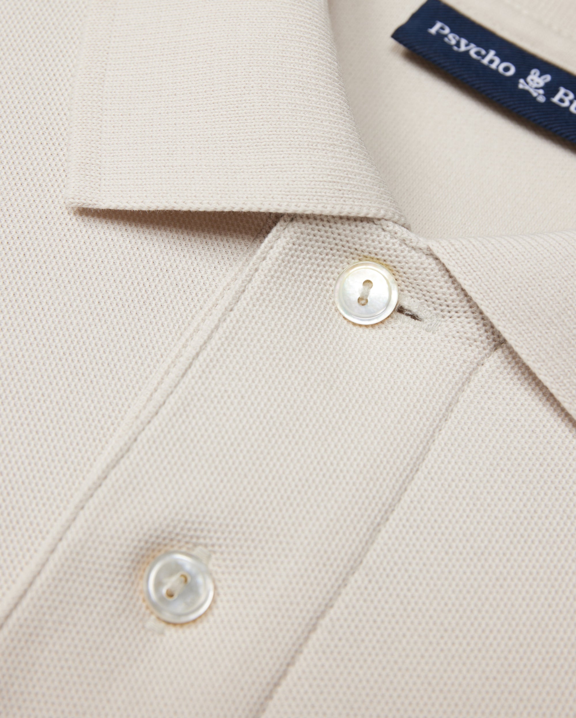 The Psycho Bunny MENS CLASSIC PIQUE POLO SHIRT - B6K001B200 in beige is crafted from luxurious Pima cotton with textured fabric. It features a collar, two white buttons, and a visible blue text label inside, embodying timeless men's fashion elegance.