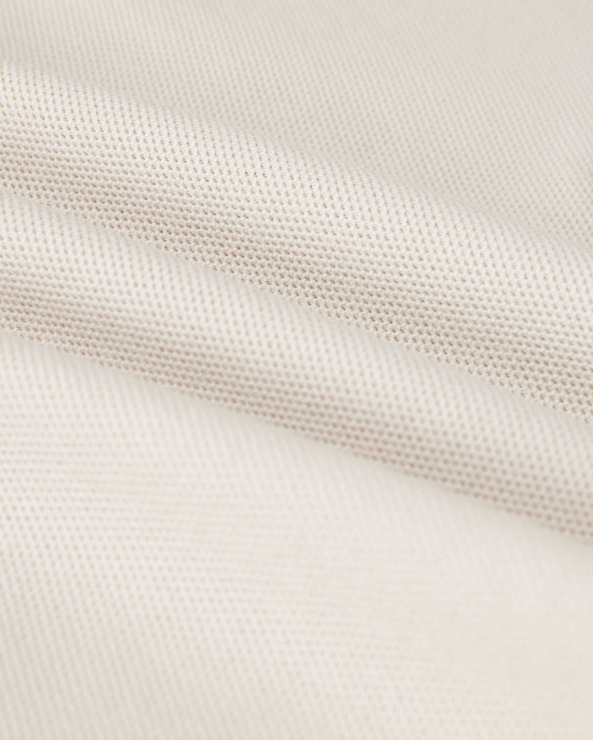 A close-up shows the Psycho Bunny MENS CLASSIC PIQUE POLO SHIRT - B6K001B200, with a textured weave pattern. The soft white fabric resembles fine Pima cotton, with subtle shadowing in the folds that exude timeless men's fashion.