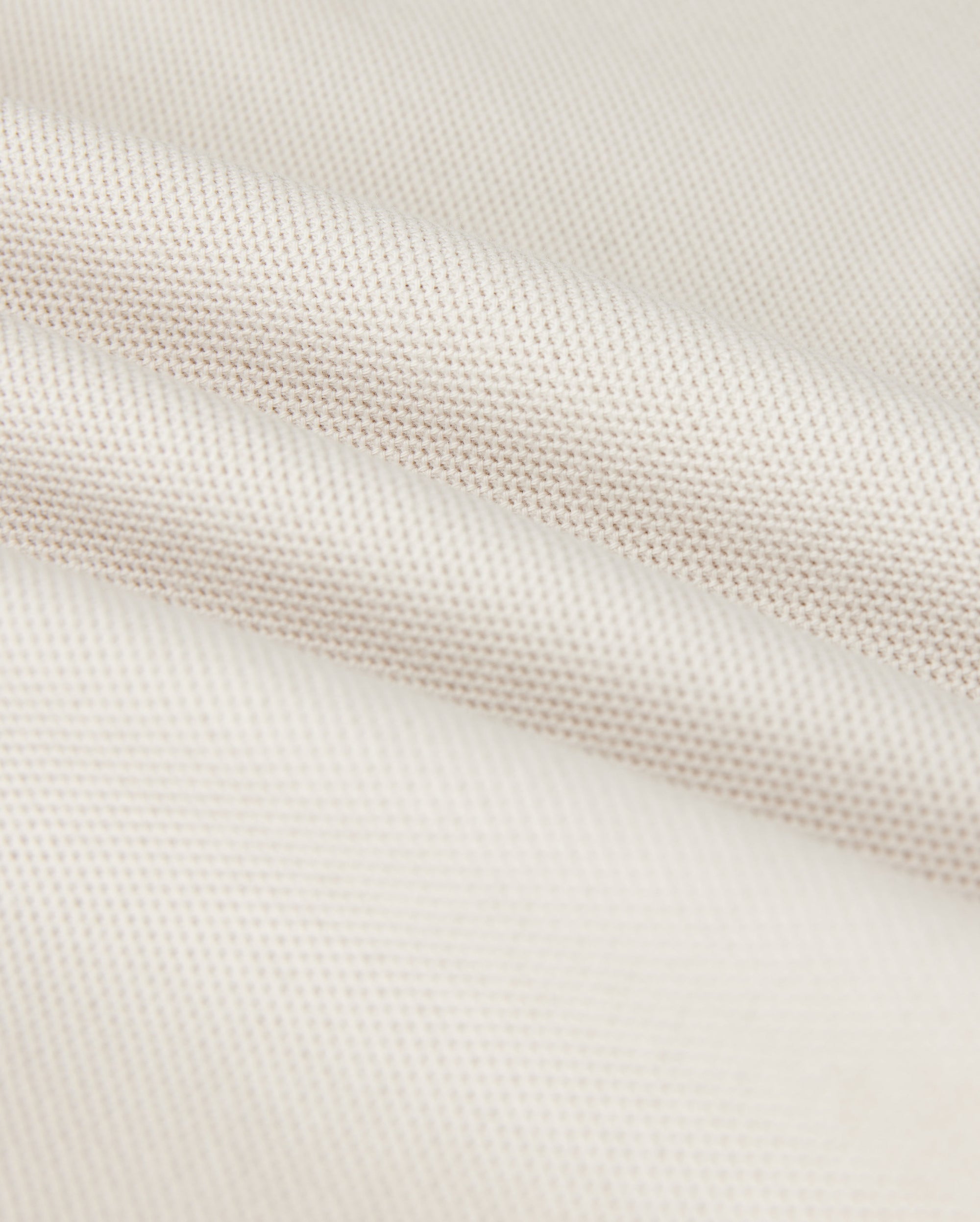 A close-up shows the Psycho Bunny MENS CLASSIC PIQUE POLO SHIRT - B6K001B200, with a textured weave pattern. The soft white fabric resembles fine Pima cotton, with subtle shadowing in the folds that exude timeless men's fashion.