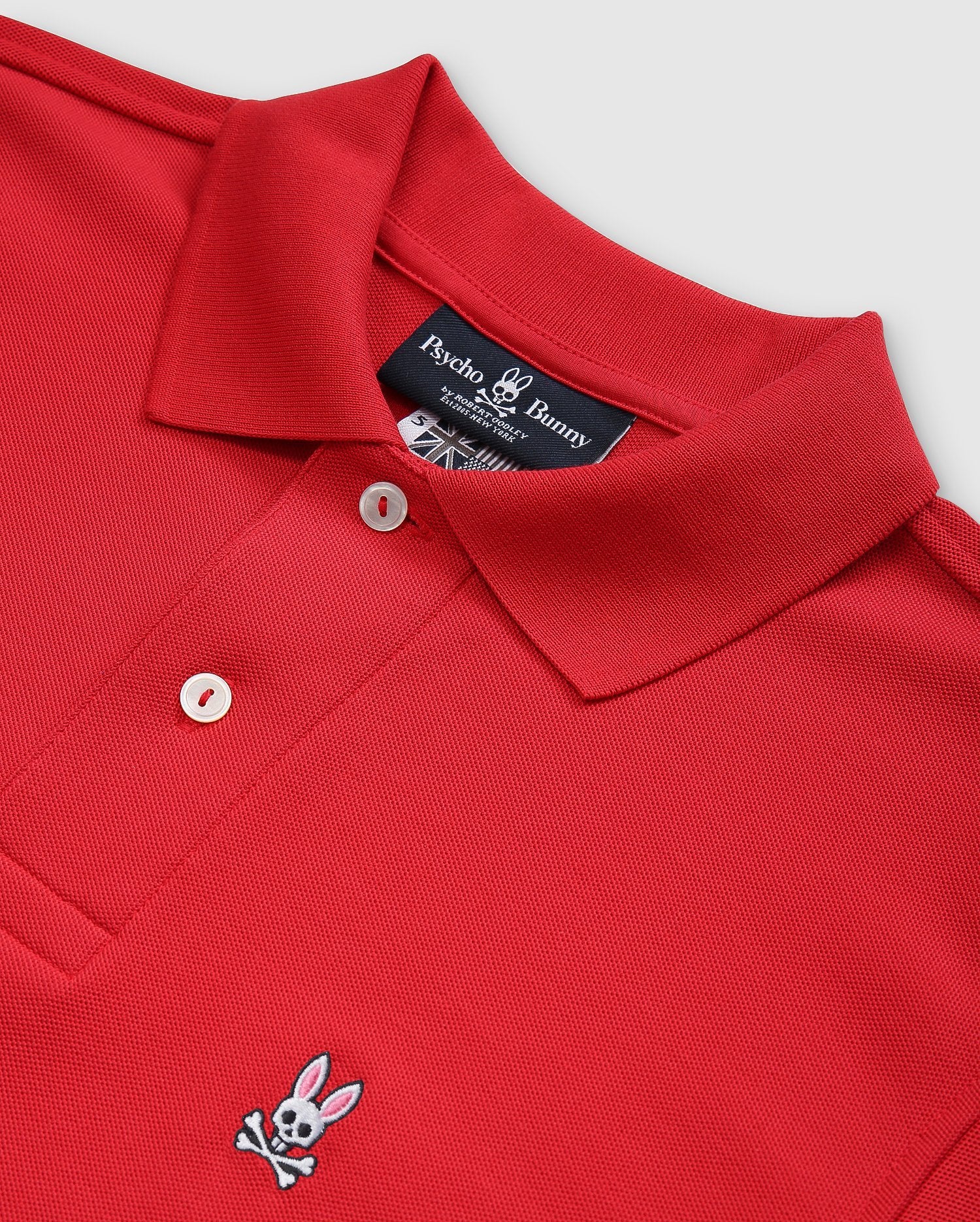 Red Psycho Bunny men's classic piqué polo shirt (B6K001CRPC), made of 100% Pima cotton, features a folded collar and two mother-of-pearl buttons. An embroidered bunny with crossbones decorates the chest. The shirt is displayed on a light gray background.
