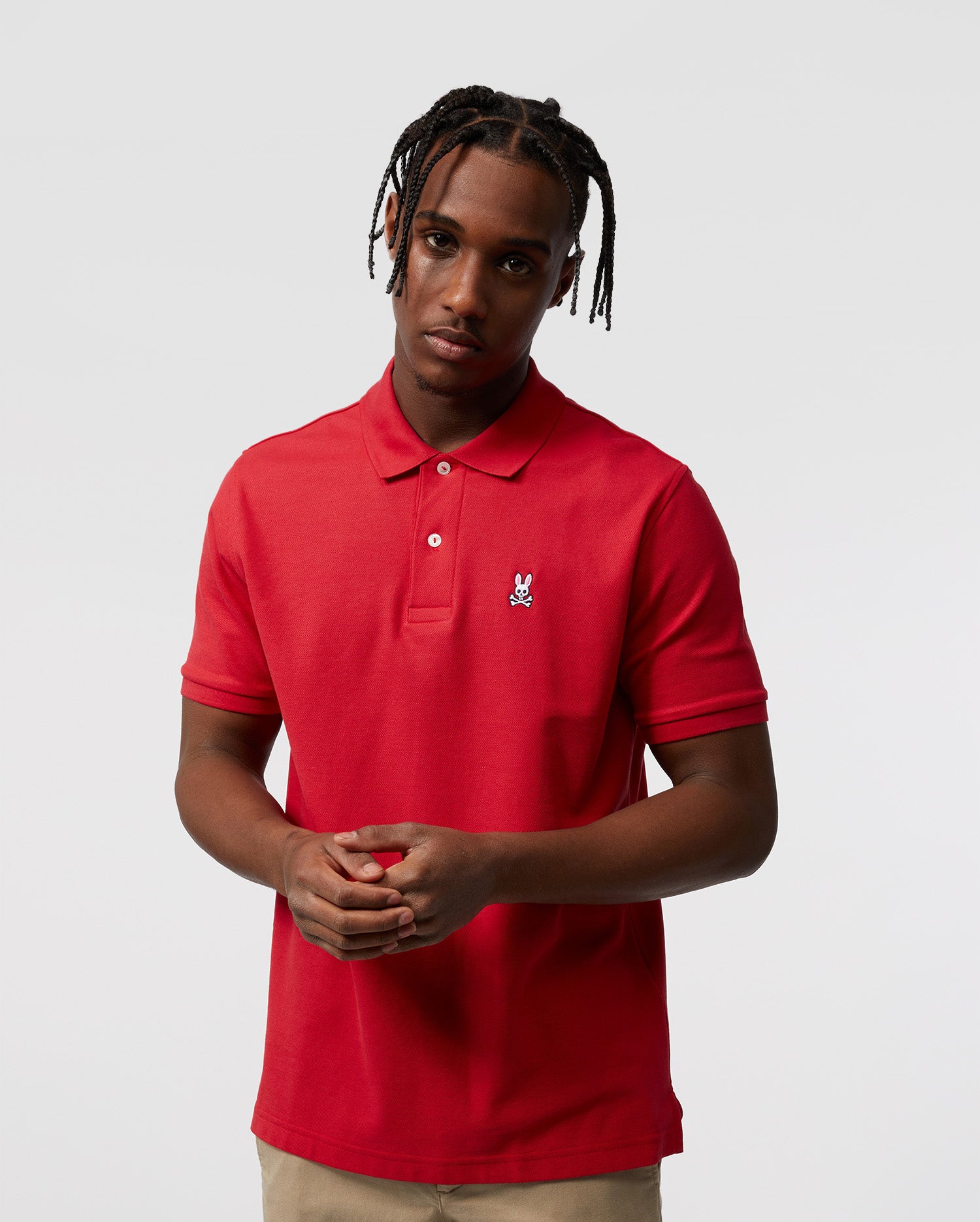 Against a plain background, a person models the Psycho Bunny Men's Classic Pique Polo Shirt (B6K001CRPC) in 100% Pima cotton with mother-of-pearl buttons, paired with beige pants. Their hair is braided, and they hold their hands together in front.