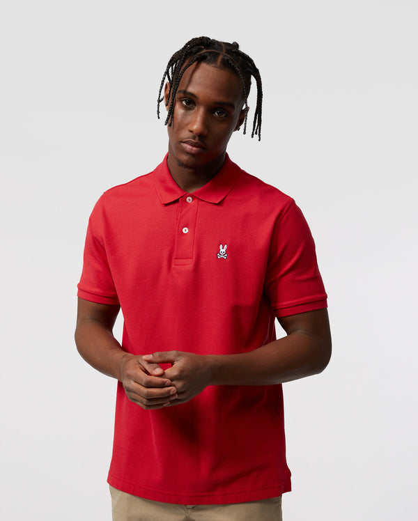 Against a plain background, a person models the Psycho Bunny Men's Classic Pique Polo Shirt (B6K001CRPC) in 100% Pima cotton with mother-of-pearl buttons, paired with beige pants. Their hair is braided, and they hold their hands together in front.