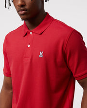 The Psycho Bunny Men's Classic Pique Polo Shirt (B6K001CRPC) is made from 100% Pima cotton with an embroidered bunny and crossbones on a red background. It has two mother-of-pearl buttons under its short sleeves against a simple white backdrop.