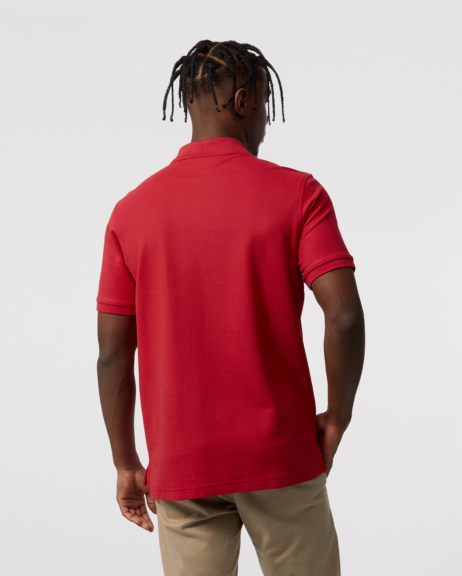 A person with braided hair stands with their back to the camera, wearing a Psycho Bunny red Men's Classic Pique Polo Shirt - B6K001CRPC made from 100% Pima cotton and beige pants, against a plain white background.