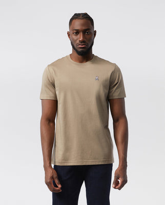 A man stands facing forward wearing a beige, 100% Pima cotton Psycho Bunny MENS CLASSIC CREW NECK TEE - B6U014CRPC with a small embroidered logo on the left chest. Sporting short hair with a styled pattern on top and a beard, he pairs his classic crew neck tee with dark jeans. The background is plain white.