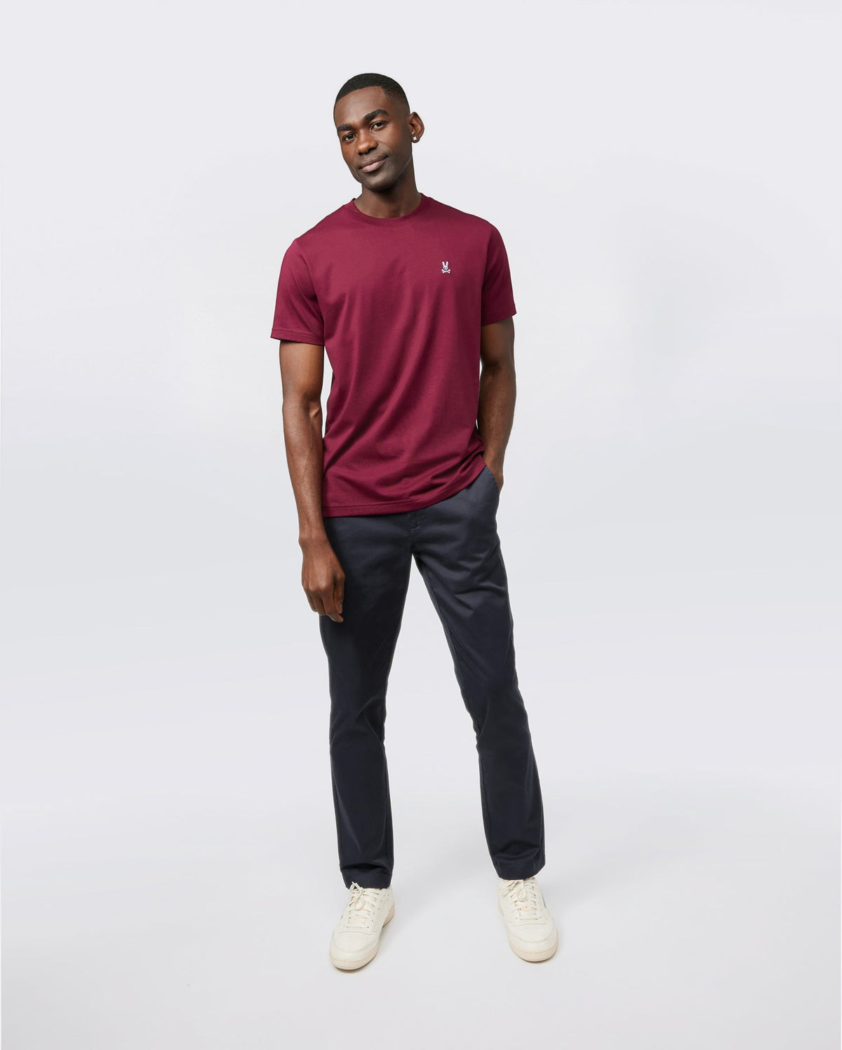 Against a plain background, a person wears a burgundy Psycho Bunny MENS CLASSIC CREW NECK TEE in ultra-soft Pima cotton. Paired with dark trousers and light sneakers, the breathable, lightweight look complements their relaxed stance as they subtly smile with hands in pockets.