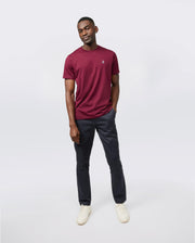 Against a plain background, a person wears a burgundy Psycho Bunny MENS CLASSIC CREW NECK TEE in ultra-soft Pima cotton. Paired with dark trousers and light sneakers, the breathable, lightweight look complements their relaxed stance as they subtly smile with hands in pockets.
