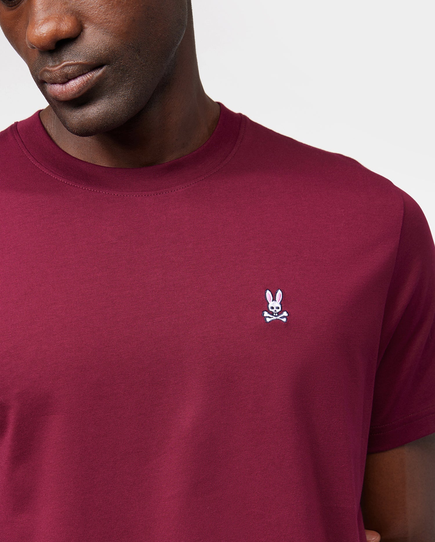 A person wears a maroon Psycho Bunny Men's Classic Crewneck Tee (B6U014Z1PC) made from ultra-soft Pima cotton. The shirt displays a small white bunny head with crossed bones logo on the left chest. The image focuses on the upper torso against a plain white background.