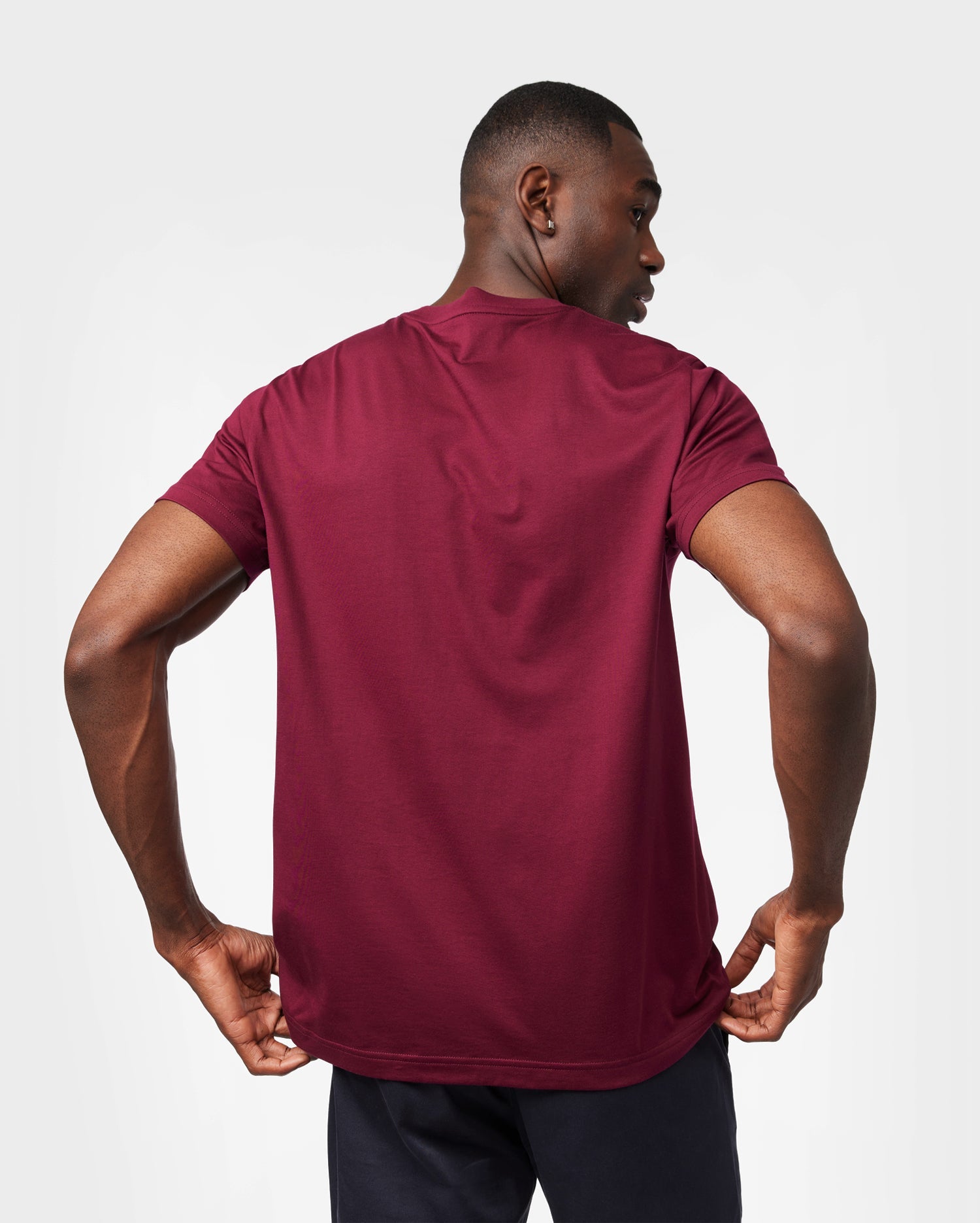 A man wears the Psycho Bunny Men's Classic Crew Neck Tee (B6U014Z1PC), a burgundy ultra-soft Pima cotton top. Paired with dark pants, he faces slightly away, adjusting his tee against a plain white background that highlights its breathable, lightweight feel.