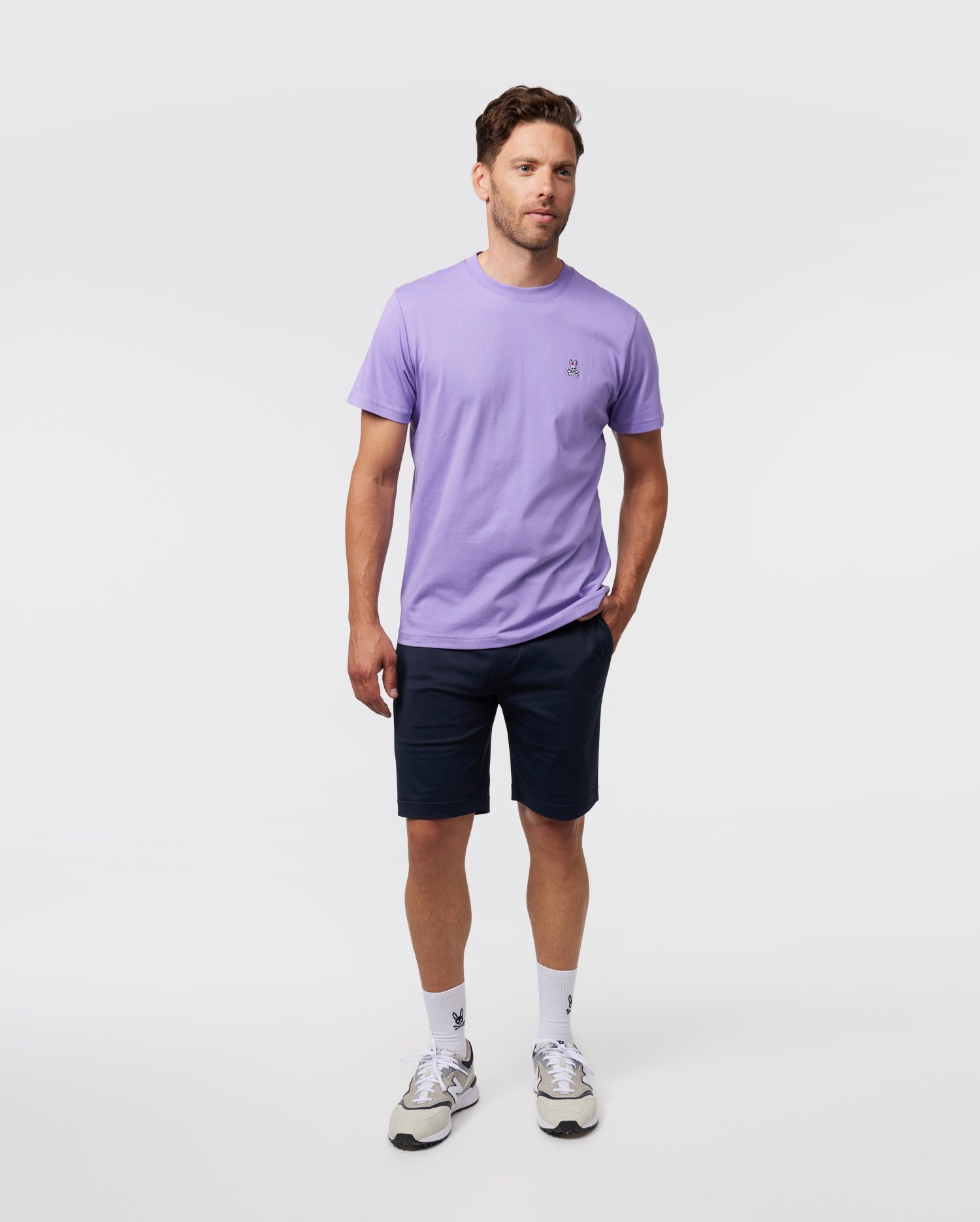 A man stands against a simple backdrop wearing a light purple MENS CLASSIC CREW NECK TEE by Psycho Bunny, dark blue shorts, white socks, and white sneakers. The soft Pima cotton tee is adorned with an embroidered Bunny logo. With short brown hair and a laid-back pose, he effortlessly exudes style.