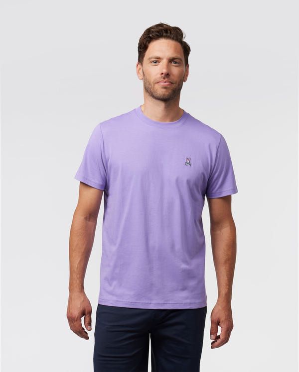 A man stands against a plain white background wearing the Psycho Bunny MENS CLASSIC CREW NECK TEE - B6U014Z1PC in a light purple shade, made from Pima cotton. The tee is adorned with an embroidered Bunny logo on the left side. He has short hair, a light beard, and is paired with dark pants.