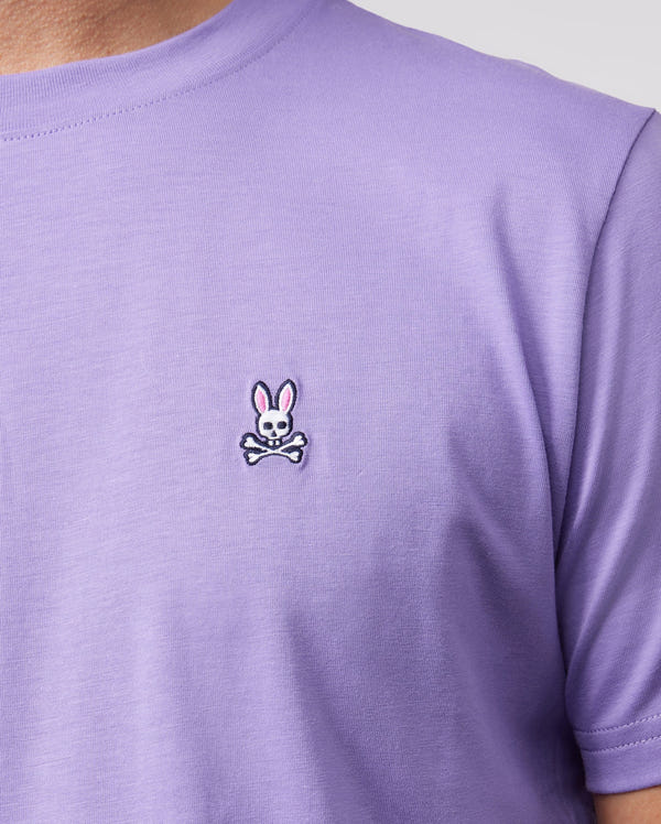 The image shows a person wearing the Psycho Bunny MENS CLASSIC CREW NECK TEE - B6U014Z1PC, crafted from luxurious Pima cotton. This lavender T-shirt is adorned with an embroidered bunny logo, featuring a rabbit head with pink ears above crossed bones in a whimsical nod to the classic skull and crossbones motif.