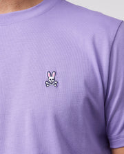 The image shows a person wearing the Psycho Bunny MENS CLASSIC CREW NECK TEE - B6U014Z1PC, crafted from luxurious Pima cotton. This lavender T-shirt is adorned with an embroidered bunny logo, featuring a rabbit head with pink ears above crossed bones in a whimsical nod to the classic skull and crossbones motif.