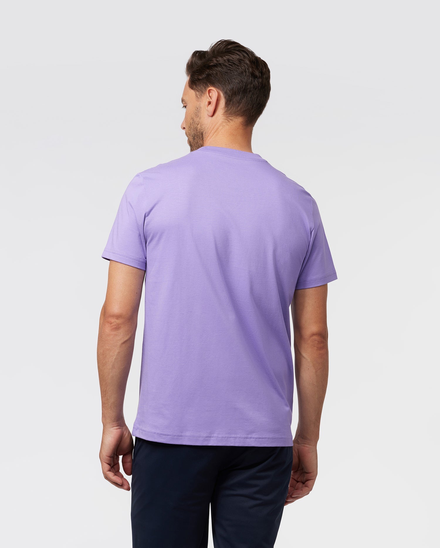 A man with short hair is wearing a Psycho Bunny MENS CLASSIC CREW NECK TEE - B6U014Z1PC in light purple Pima cotton paired with dark pants. He stands with his back to the camera against a plain light gray background, highlighting the comfort and quality of his attire.