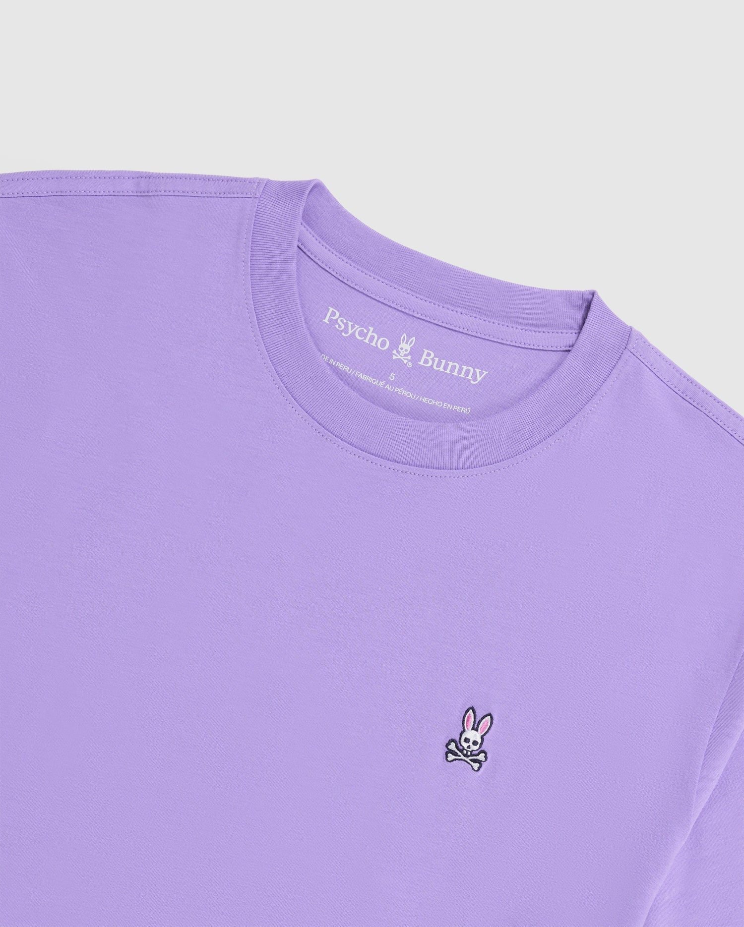 This classic crew neck tee, known as the MENS CLASSIC CREW NECK TEE - B6U014Z1PC by Psycho Bunny, comes in a lavender shade and is made from soft Pima cotton. It features a small embroidered Bunny logo with crossbones on the left side and includes an interior neck label that displays the "Psycho Bunny" brand name against a simple, light background.