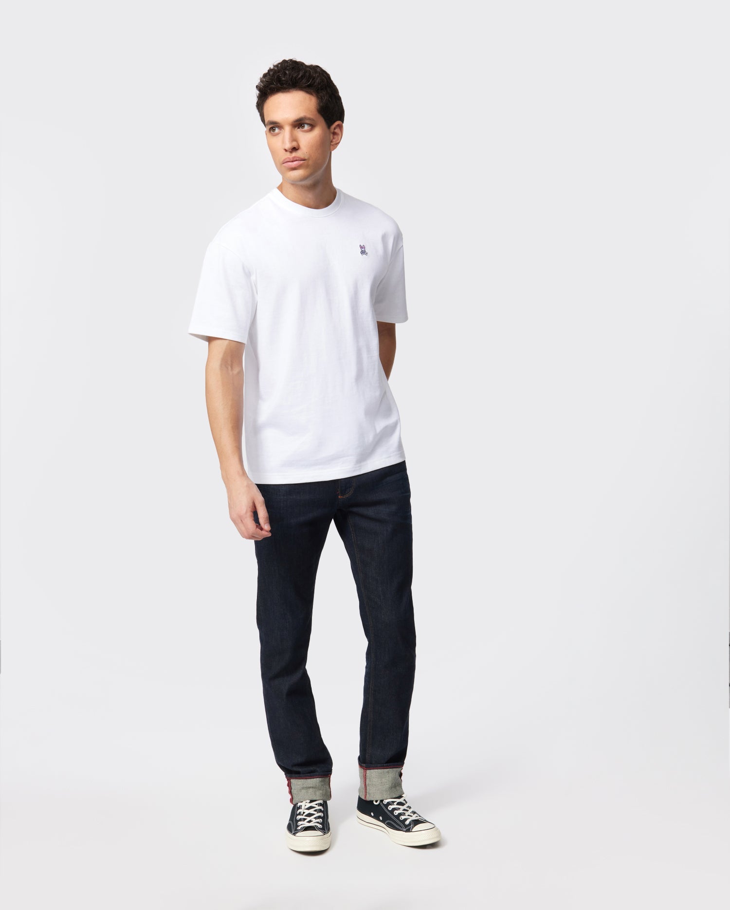A man stands against a plain white background. He is wearing a plain white Psycho Bunny Peruvian Pima cotton crew neck tee (MENS OVERSIZED CLASSIC CREW NECK TEE - B6U240W1PC), dark blue jeans with rolled cuffs, and black sneakers with white laces and soles. His hands are resting by his sides, and he is looking slightly to his left.
