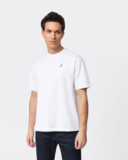 A person with short dark hair is wearing a Psycho Bunny MENS OVERSIZED CLASSIC CREW NECK TEE - B6U240W1PC, made from Peruvian Pima cotton and featuring a small embroidered design on the left chest, along with dark blue jeans. They are standing against a white background and looking slightly to the side.