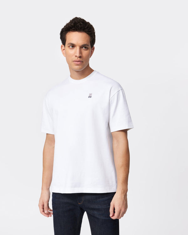 A person with short dark hair is wearing a Psycho Bunny MENS OVERSIZED CLASSIC CREW NECK TEE - B6U240W1PC, made from Peruvian Pima cotton and featuring a small embroidered design on the left chest, along with dark blue jeans. They are standing against a white background and looking slightly to the side.