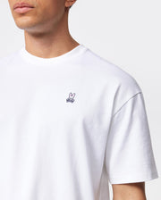 A person wearing a plain white Psycho Bunny MENS OVERSIZED CLASSIC CREW NECK TEE (model B6U240W1PC) made of Peruvian Pima cotton with a small embroidered pink rabbit logo on the left chest. The background is plain and gray. The image is cropped to show only the upper torso and part of the person's face.
