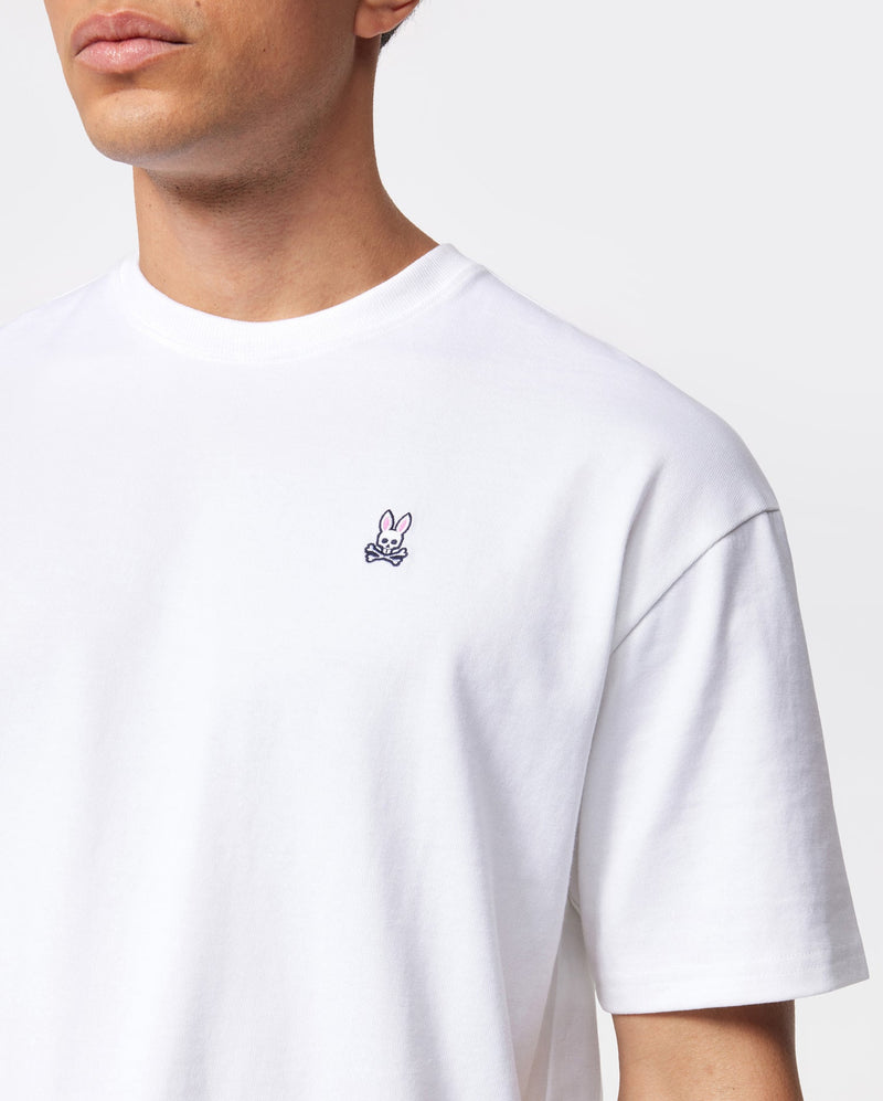 A person wearing a plain white Psycho Bunny MENS OVERSIZED CLASSIC CREW NECK TEE (model B6U240W1PC) made of Peruvian Pima cotton with a small embroidered pink rabbit logo on the left chest. The background is plain and gray. The image is cropped to show only the upper torso and part of the person's face.