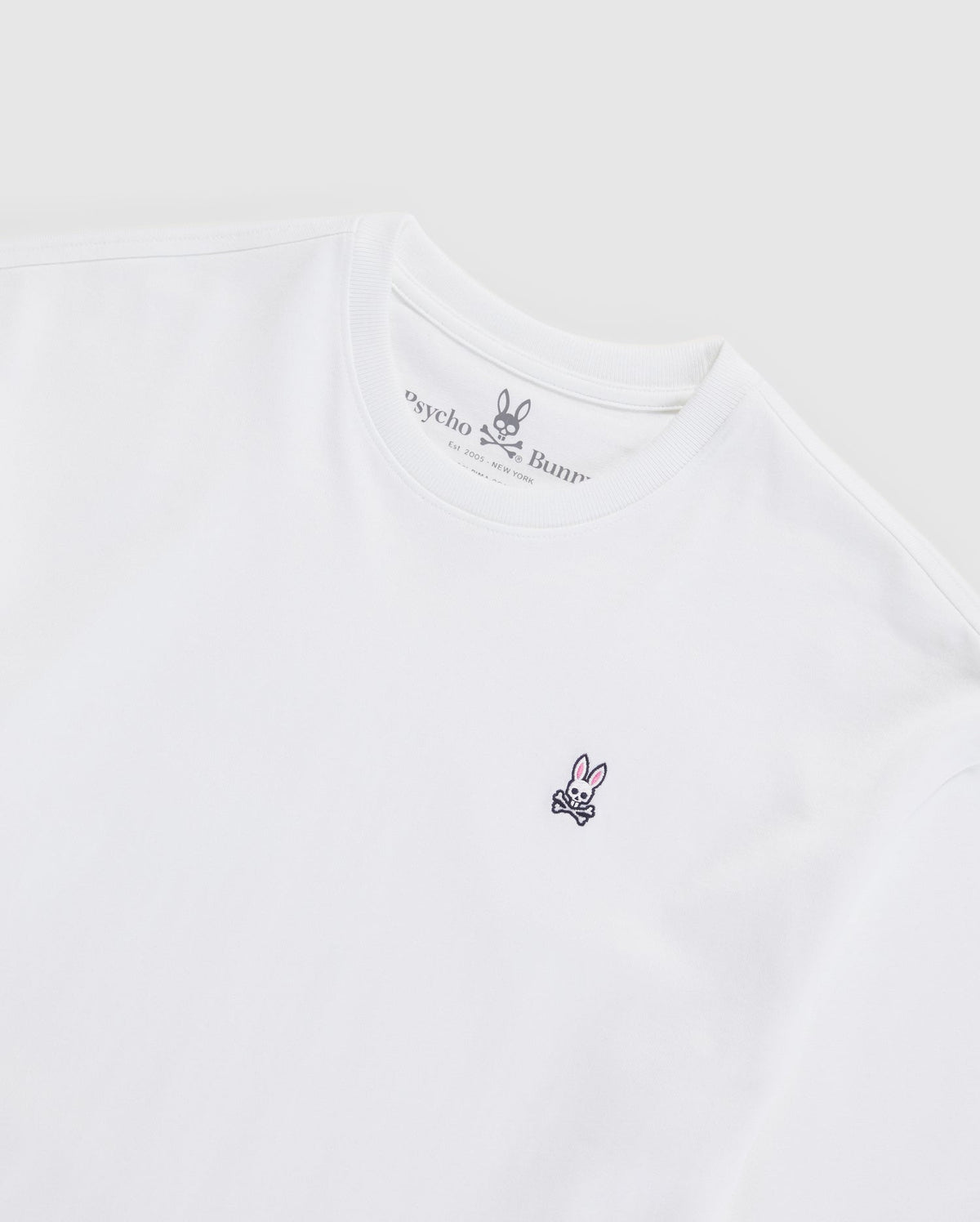 The MENS OVERSIZED CLASSIC CREW NECK TEE - B6U240W1PC by Psycho Bunny is a plain white tee made from luxurious Peruvian Pima cotton. It features a small embroidered logo on the left chest depicting a stylized bunny face and crossbones. The brand name "Psycho Bunny" is visible inside the neckline, set against a light gray background.