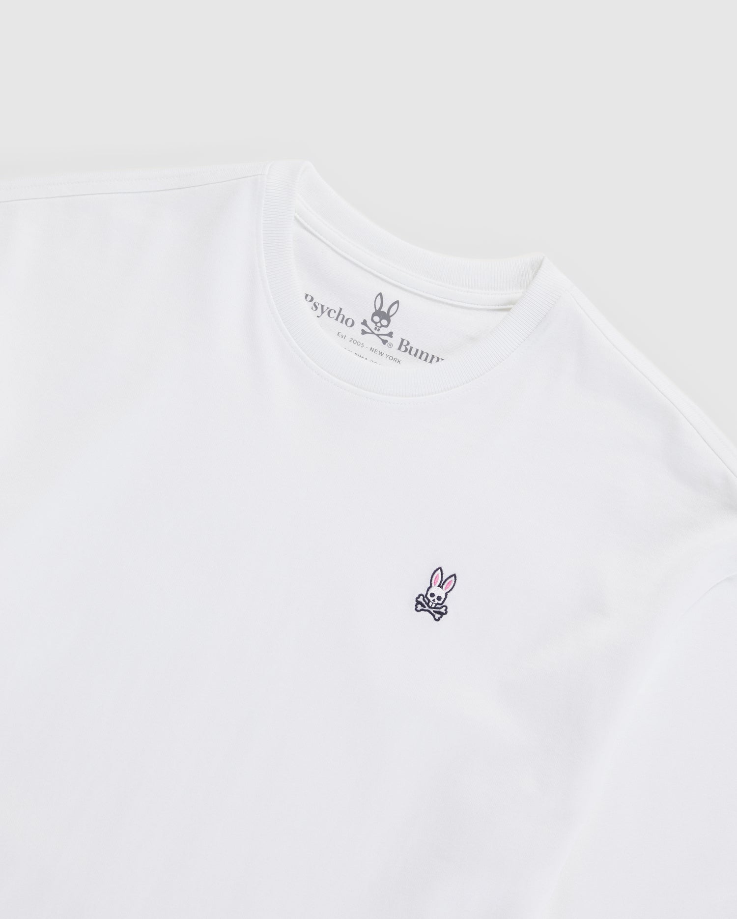 The MENS OVERSIZED CLASSIC CREW NECK TEE - B6U240W1PC by Psycho Bunny is a plain white tee made from luxurious Peruvian Pima cotton. It features a small embroidered logo on the left chest depicting a stylized bunny face and crossbones. The brand name "Psycho Bunny" is visible inside the neckline, set against a light gray background.