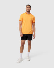 A man stands against a plain backdrop, wearing an orange T-shirt with a small chest logo, black MENS MALTA HYDROCHROMIC SWIM TRUNK - B6W321B2SW from Psycho Bunny, white socks, and white sneakers. He is smiling and has a relaxed posture with one hand by his side and the other slightly extended.